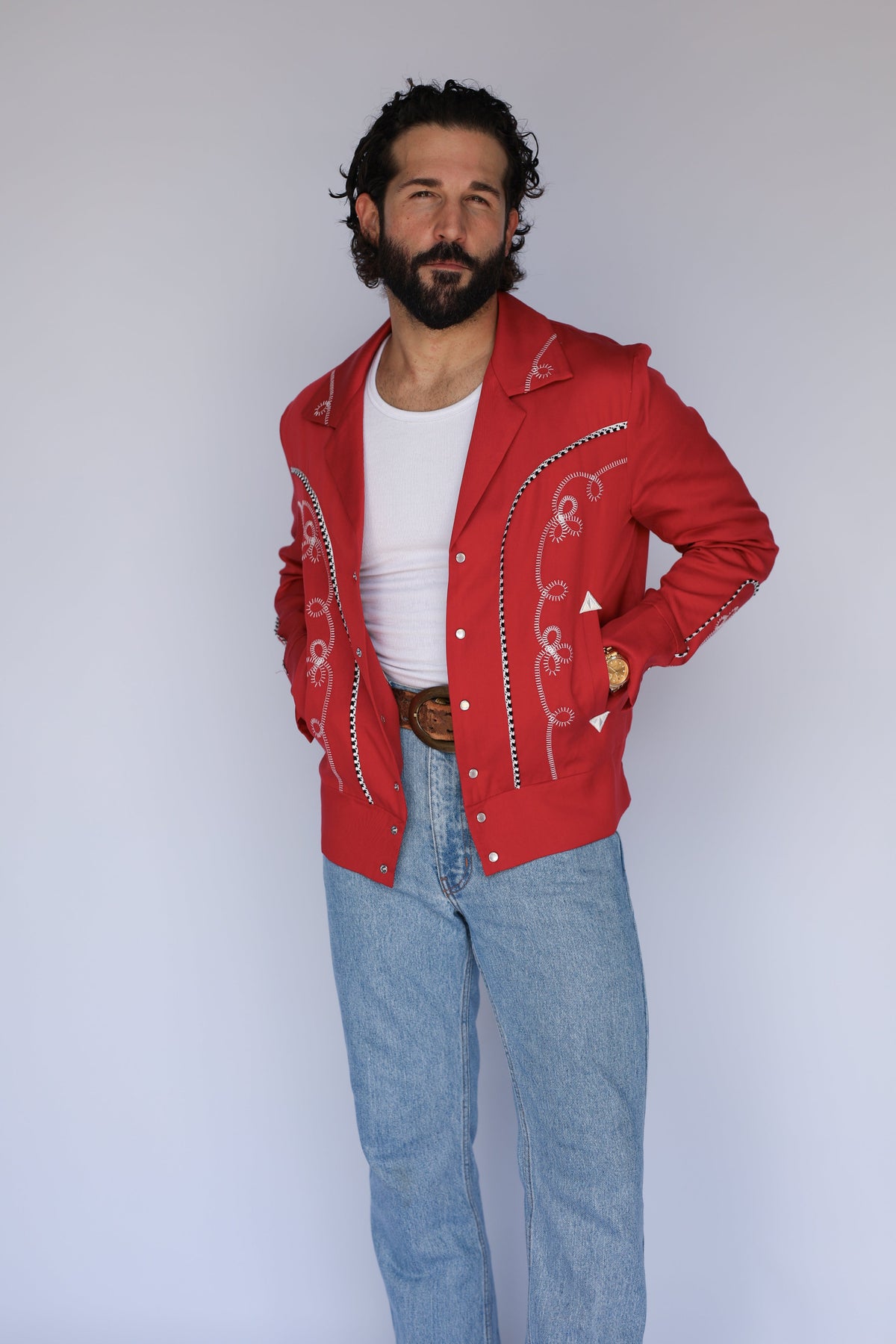 A man wearing a red western-style jacket with white embroidery, a white t-shirt, and light blue jeans, standing against a plain light gray background.