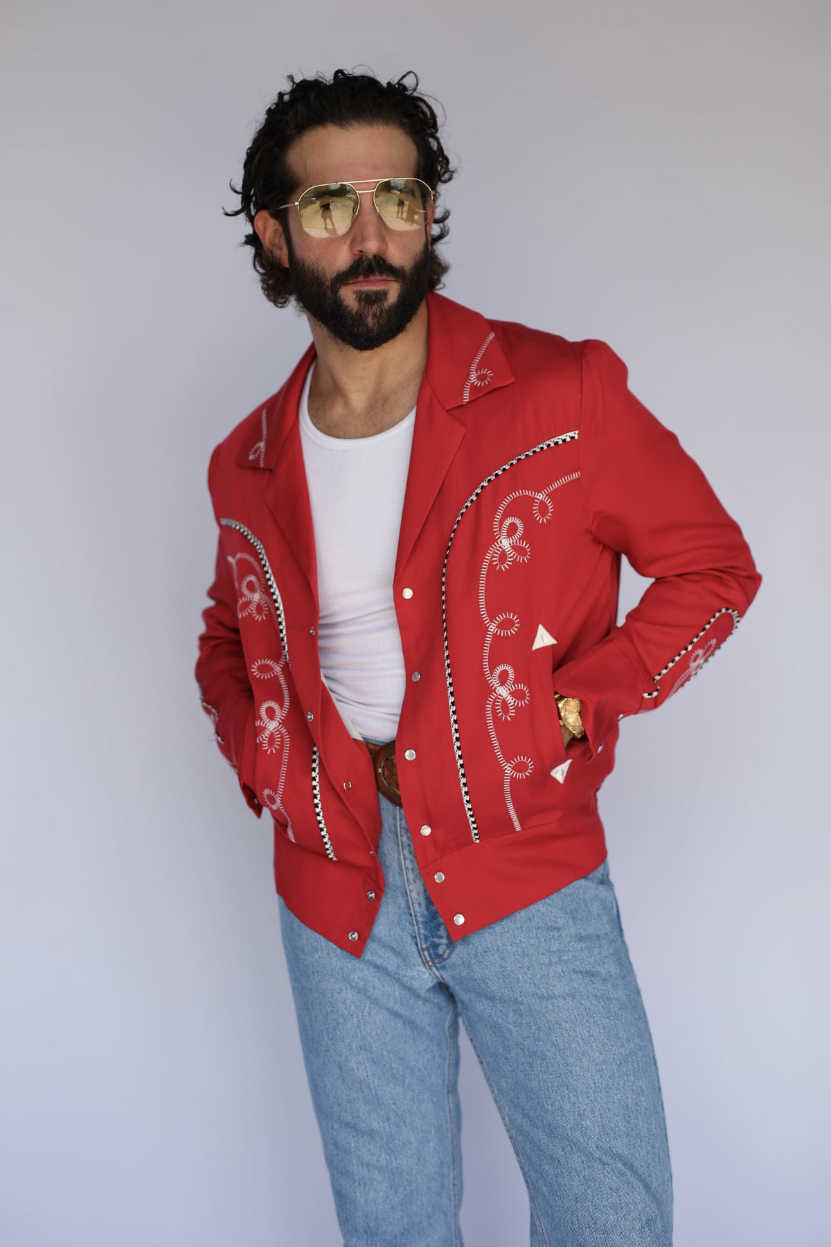A man wearing a bright red western-style jacket with white embroidery and silver trim, styled with a white t-shirt and light blue jeans against a plain light gray background.