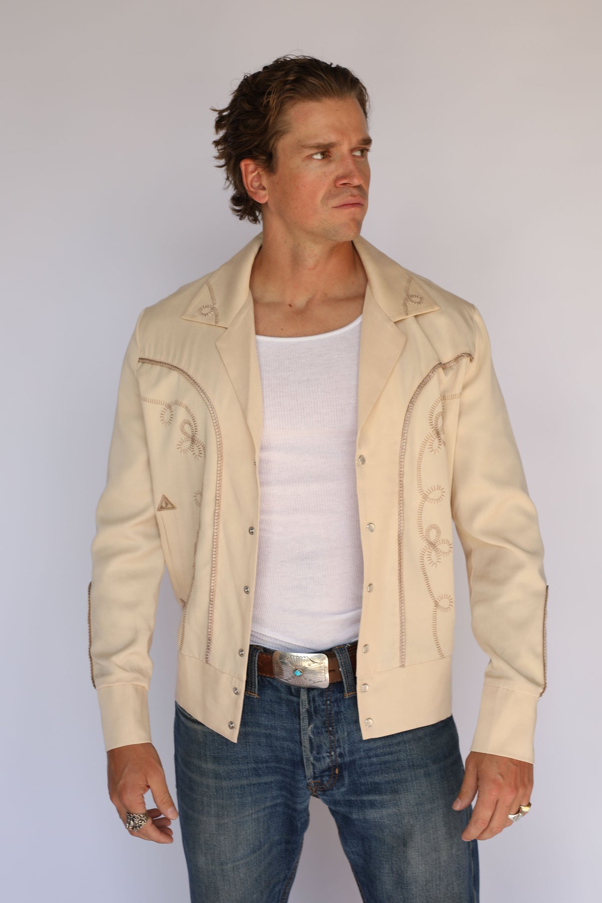A man wearing a cream western-style leather jacket with embroidered details, a white tank top, and blue jeans, standing against a plain light gray background.