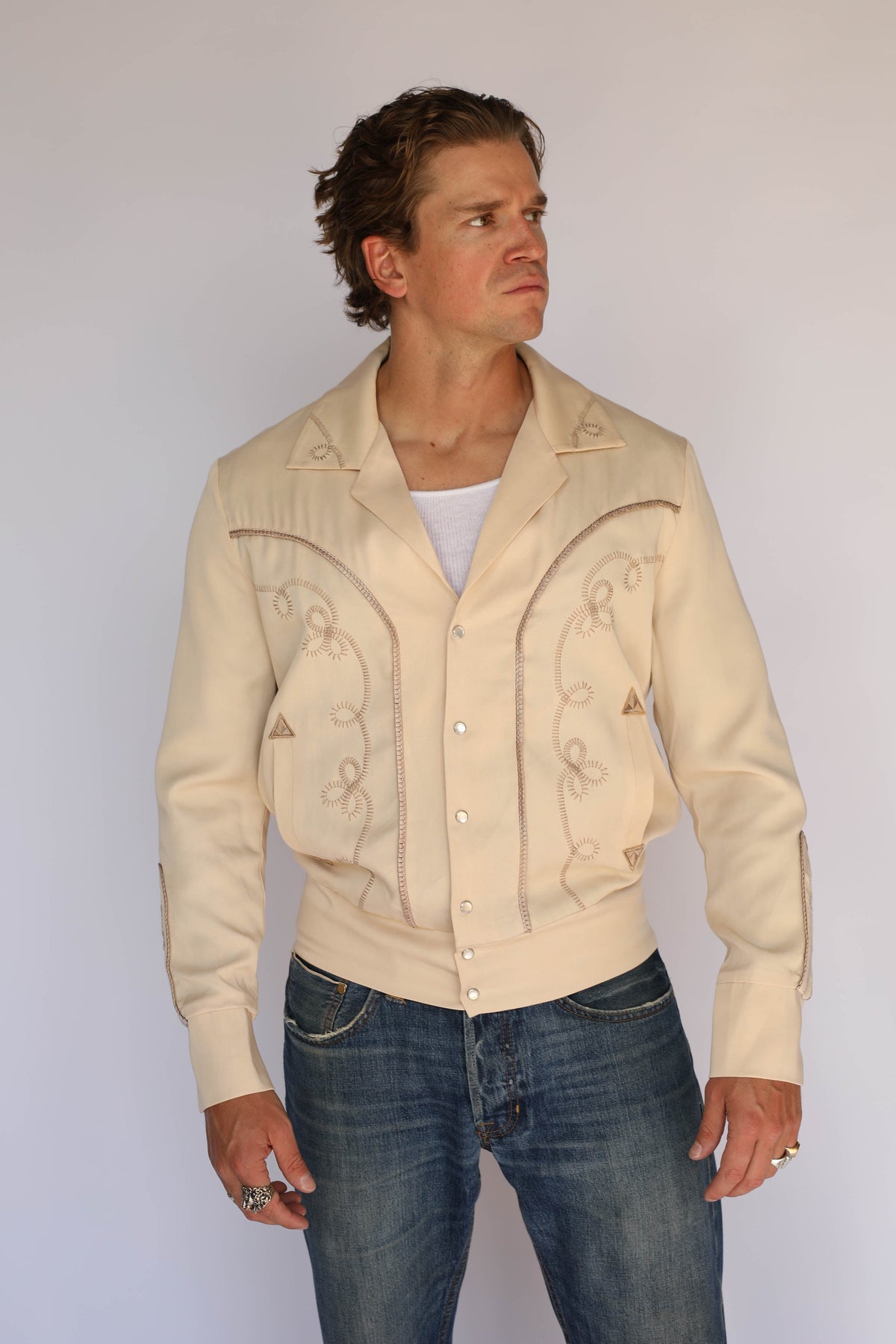 A man wearing a cream H Bar C vintage inspired western-style jacket with embroidered details, a white undershirt, and blue jeans, standing against a plain light gray background.