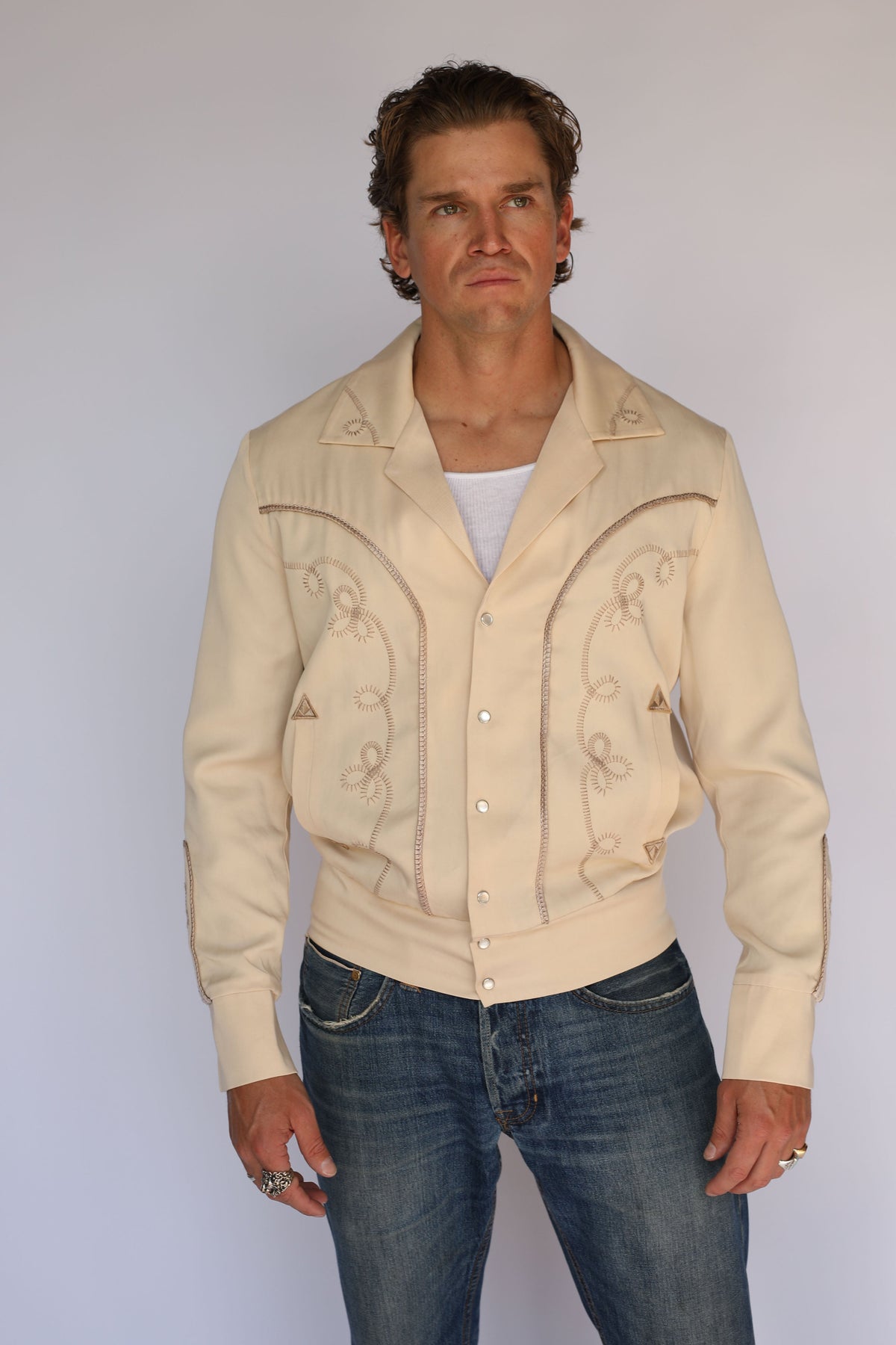 A man wearing a cream H Bar C vintage inspired western-style jacket with embroidered details, a white undershirt, and blue jeans, standing against a plain light gray background.