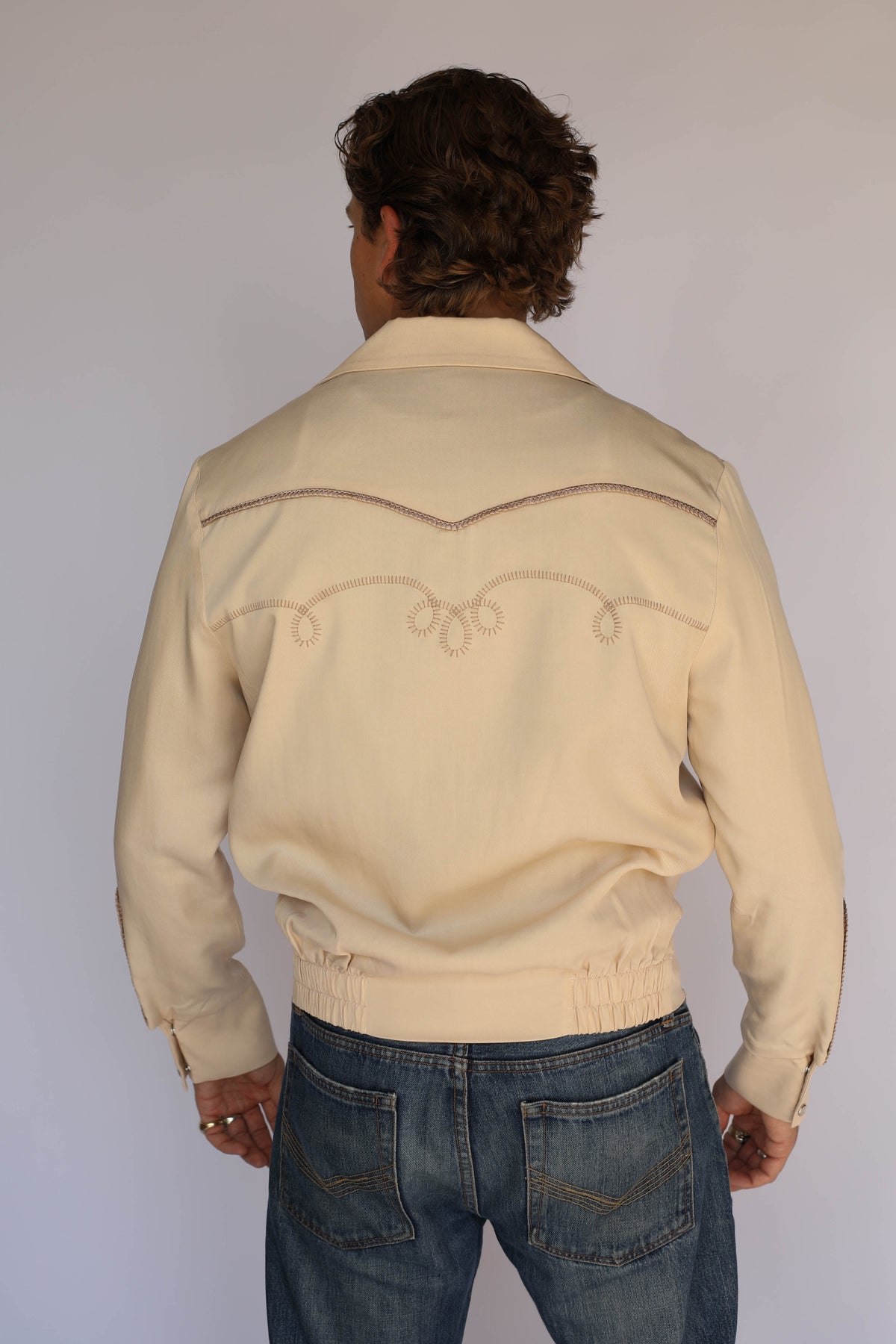 A man wearing a cream H Bar C vintage inspired western-style jacket with embroidered details, a white undershirt, and blue jeans, standing against a plain light gray background.