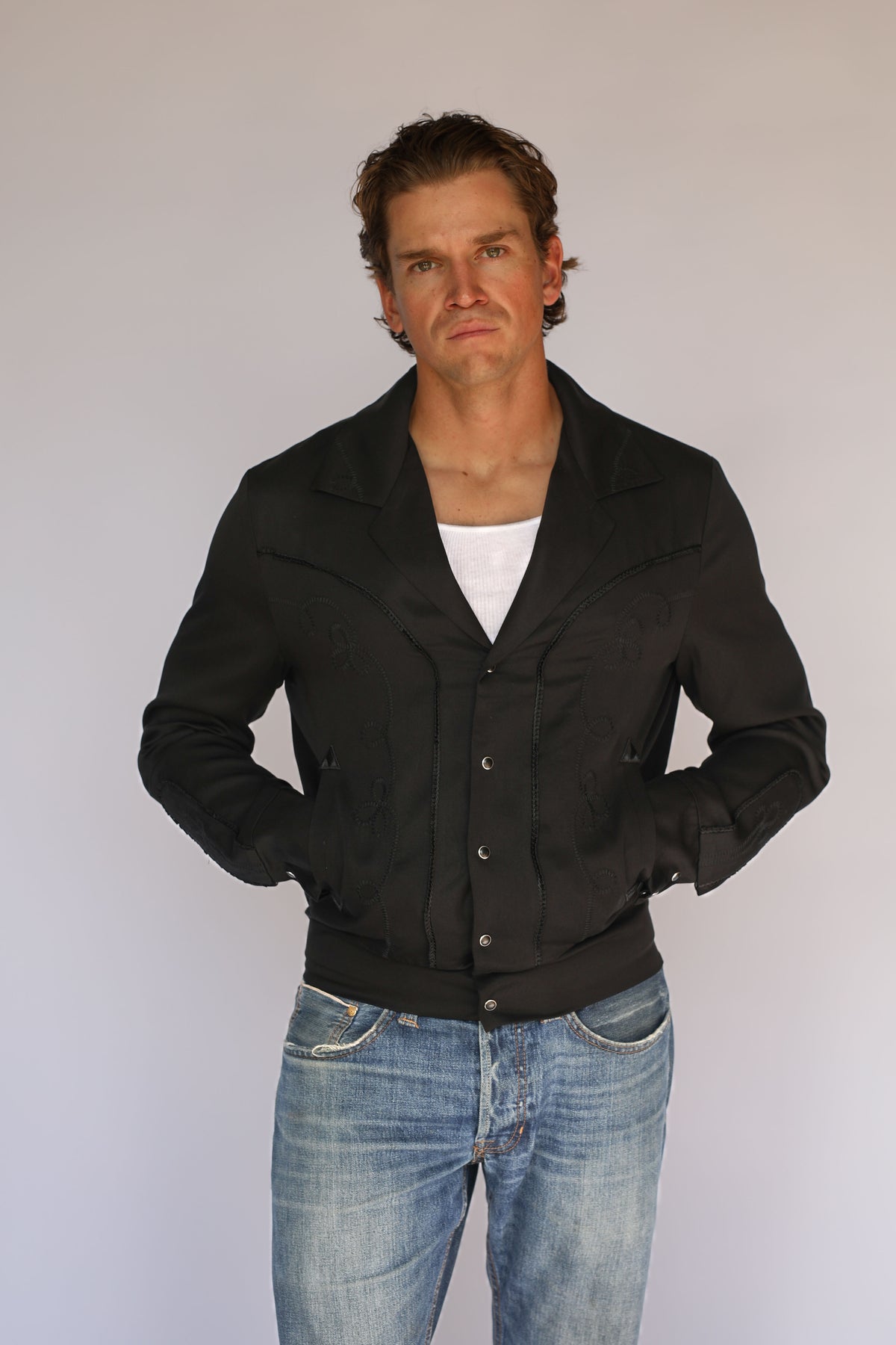 A man wearing a black H Bar C vintage inspired western-style snap-button jacket over a white t-shirt and blue jeans, standing against a plain light gray background.