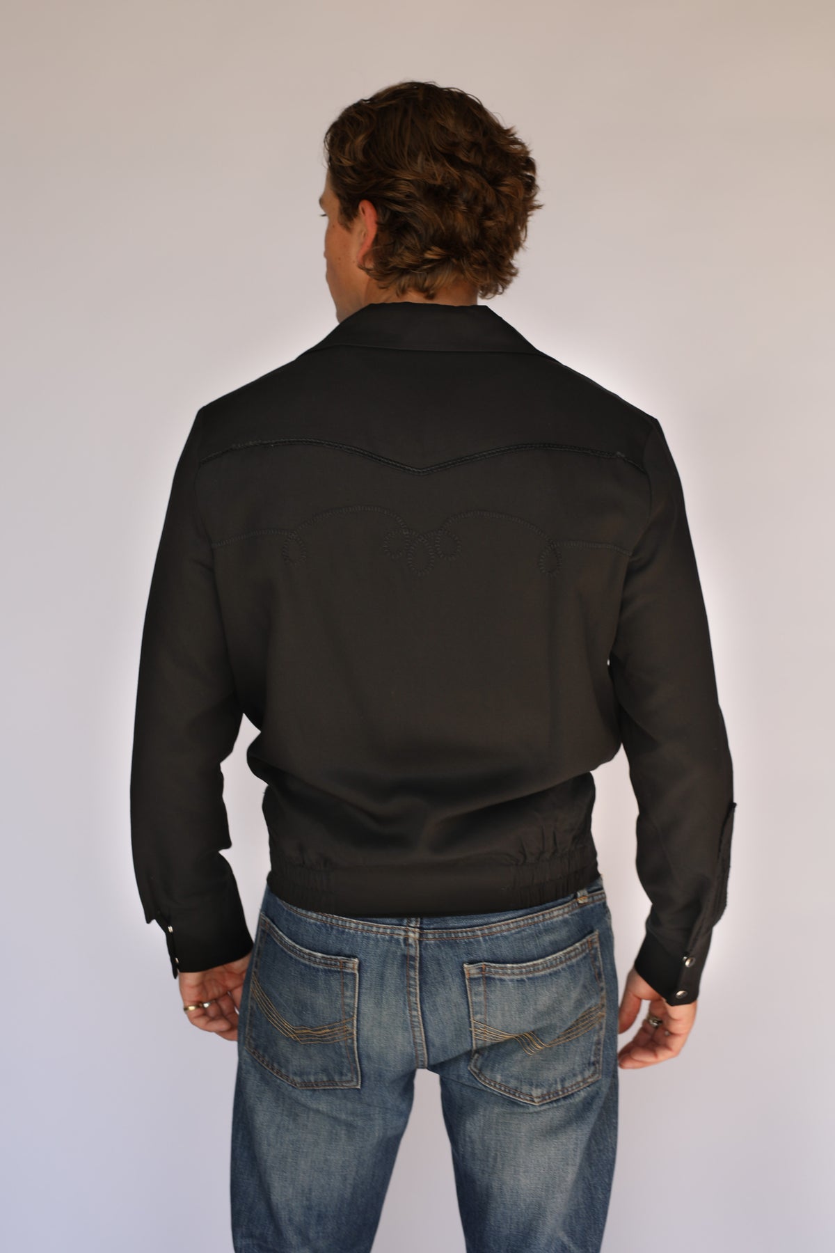 A man wearing a black H Bar C vintage inspired western-style snap-button jacket over a white t-shirt and blue jeans, standing against a plain light gray background.