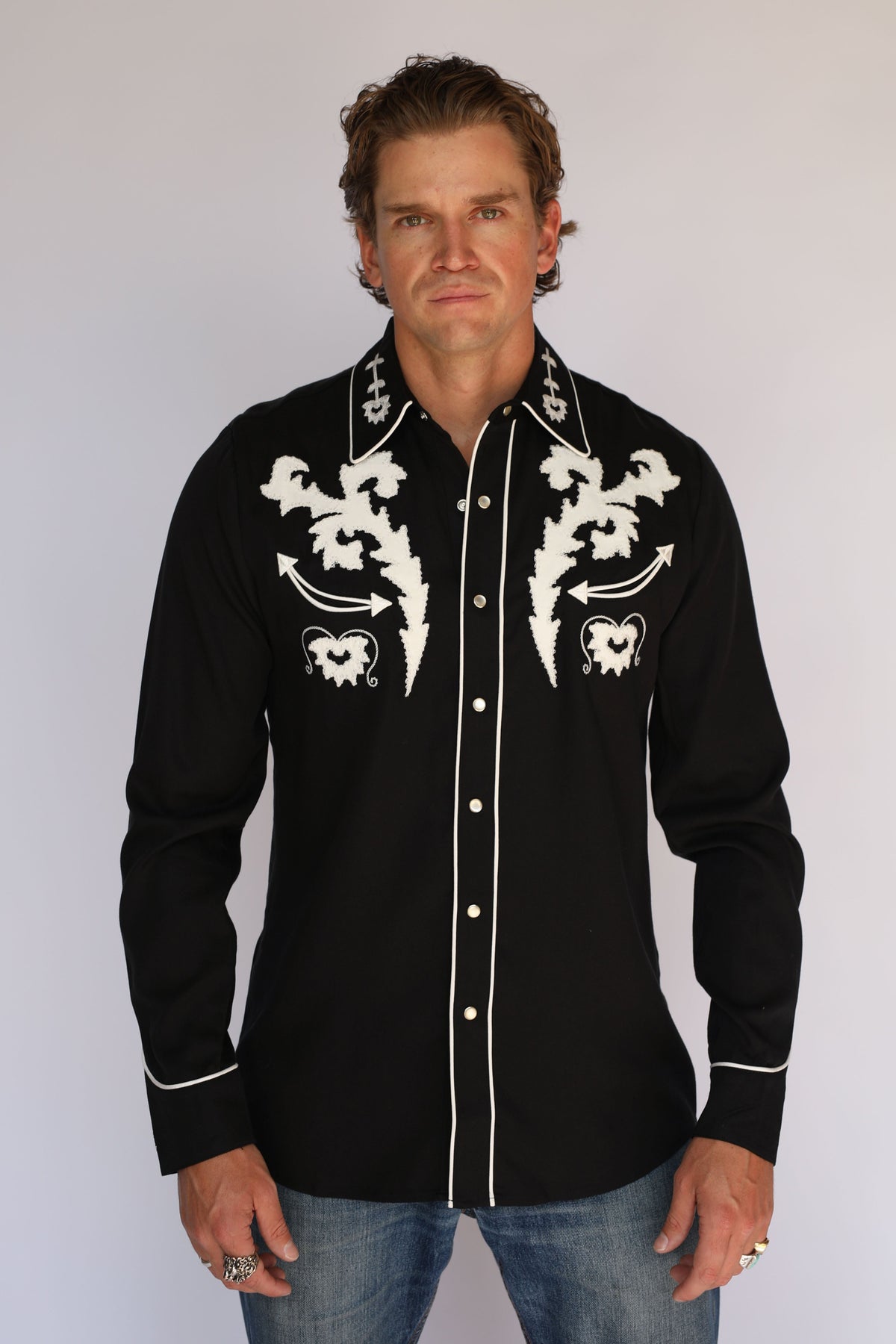 A man wearing an H Bar C black long-sleeve western shirt with white embroidered patterns on the chest and collar, paired with blue jeans and a brown belt, standing against a light gray background.