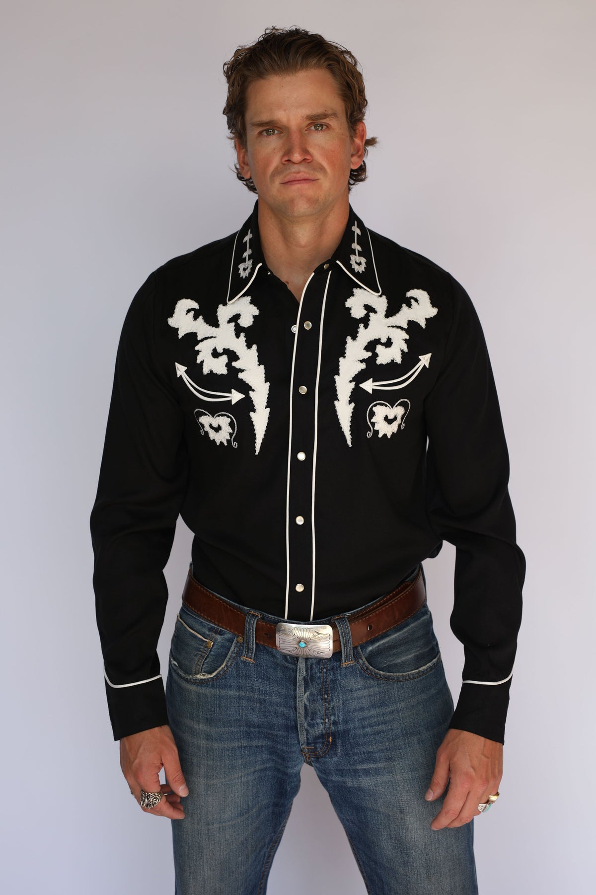 A man wearing an H Bar C black long-sleeve western shirt with white embroidered patterns on the chest and collar, paired with blue jeans and a brown belt, standing against a light gray background.