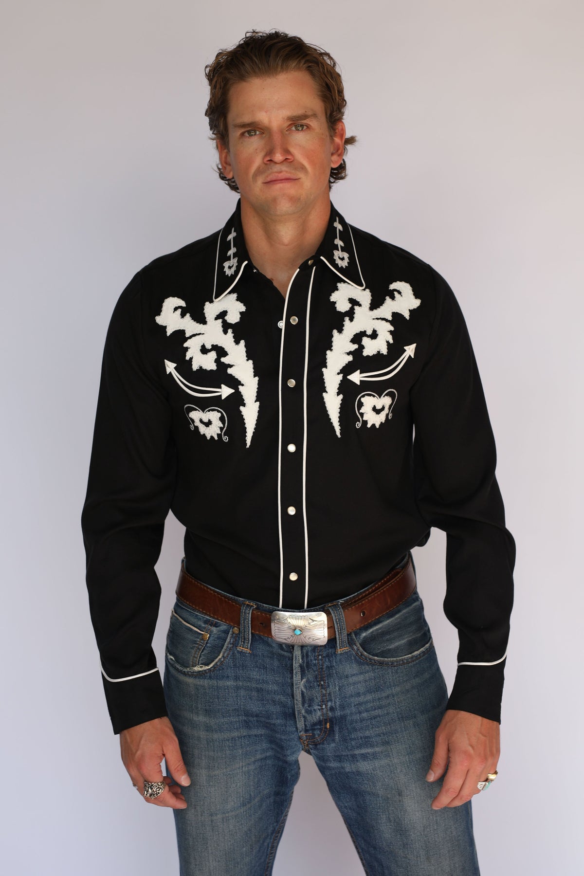 A man wearing an H Bar C black long-sleeve western shirt with white embroidered patterns on the chest and collar, paired with blue jeans and a brown belt, standing against a light gray background.