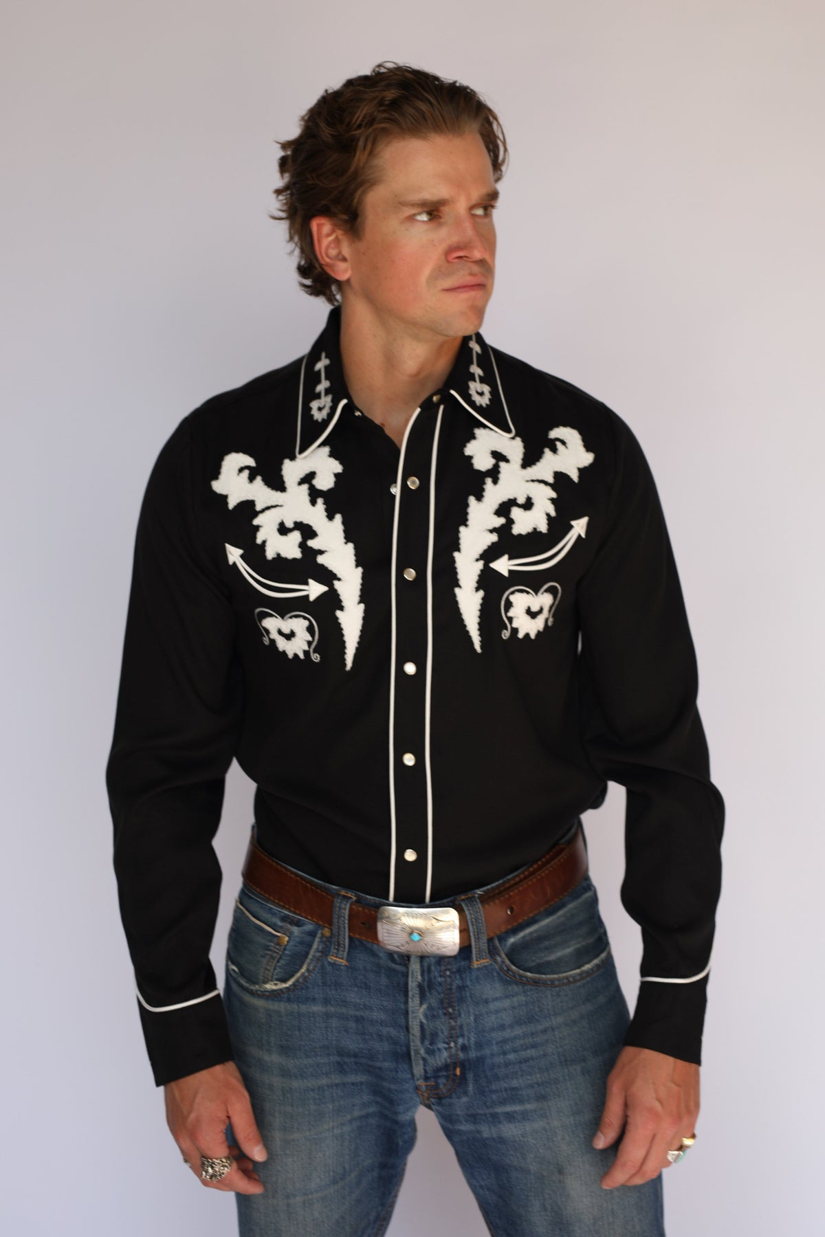A man wearing an H Bar C black long-sleeve western shirt with white embroidered patterns on the chest and collar, paired with blue jeans and a brown belt, standing against a light gray background.