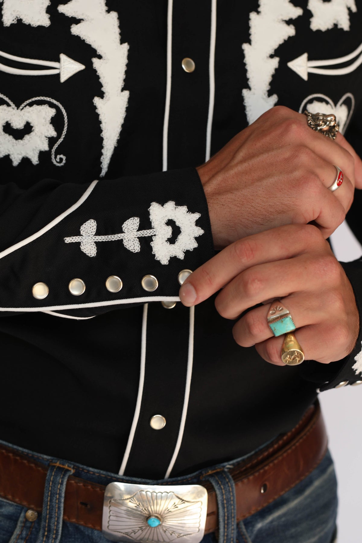 A man wearing an H Bar C black long-sleeve western shirt with white embroidered patterns on the chest and collar, paired with blue jeans and a brown belt, standing against a light gray background.