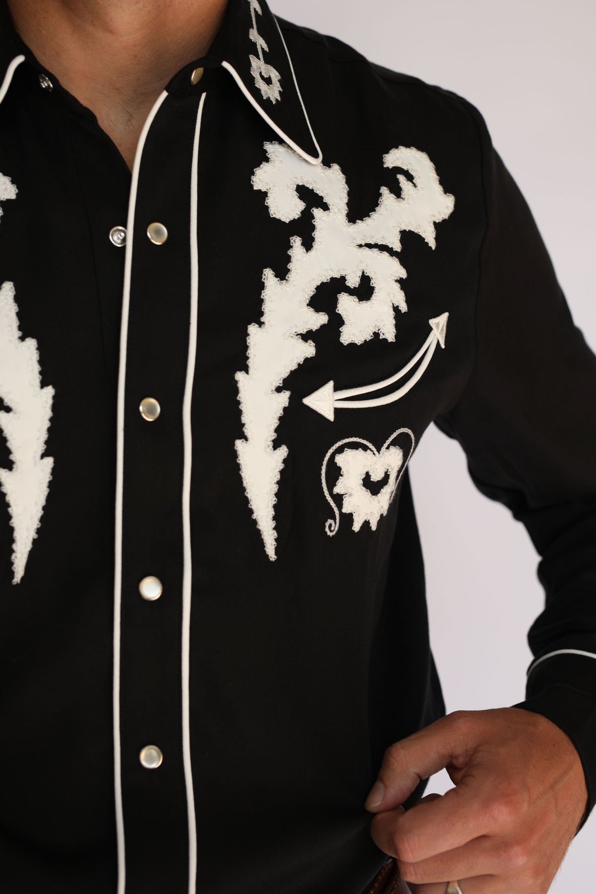 A man wearing an H Bar C black long-sleeve western shirt with white embroidered patterns on the chest and collar, paired with blue jeans and a brown belt, standing against a light gray background.