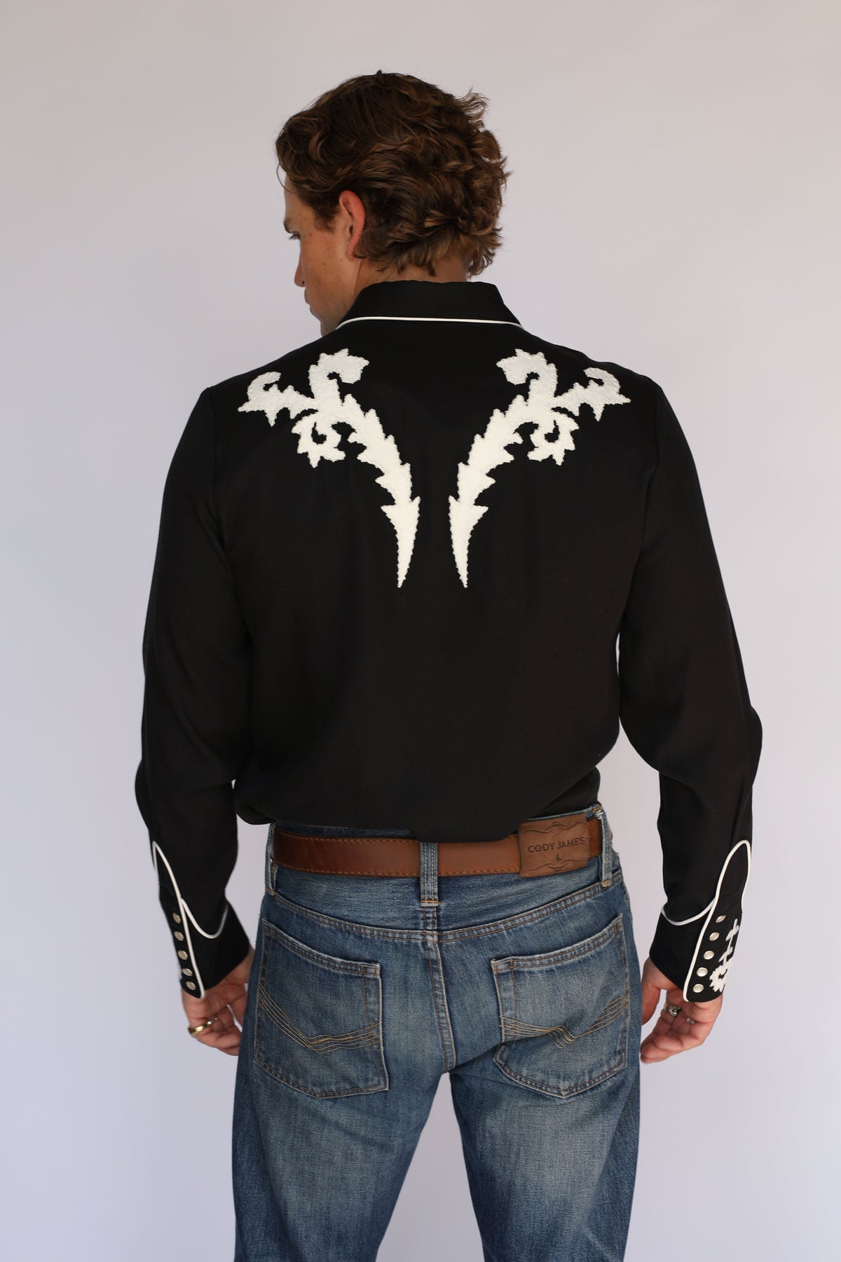 A man wearing an H Bar C black long-sleeve western shirt with white embroidered patterns on the chest and collar, paired with blue jeans and a brown belt, standing against a light gray background.