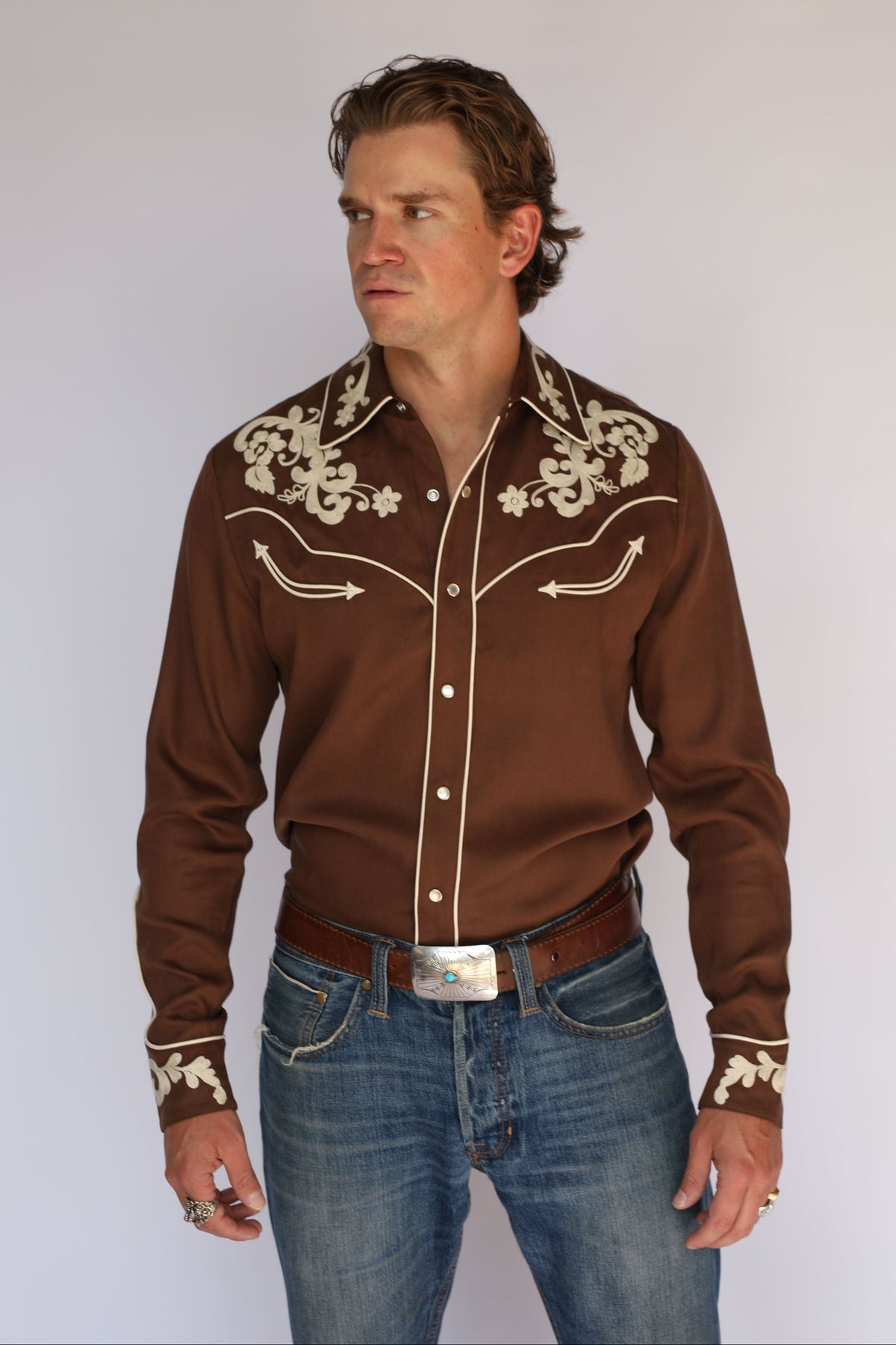 A man wearing an H Bar C Durango brown shirt, vintage inspired western wear with cream embroidery on the collar, chest, and cuffs, paired with blue jeans and a silver belt buckle.