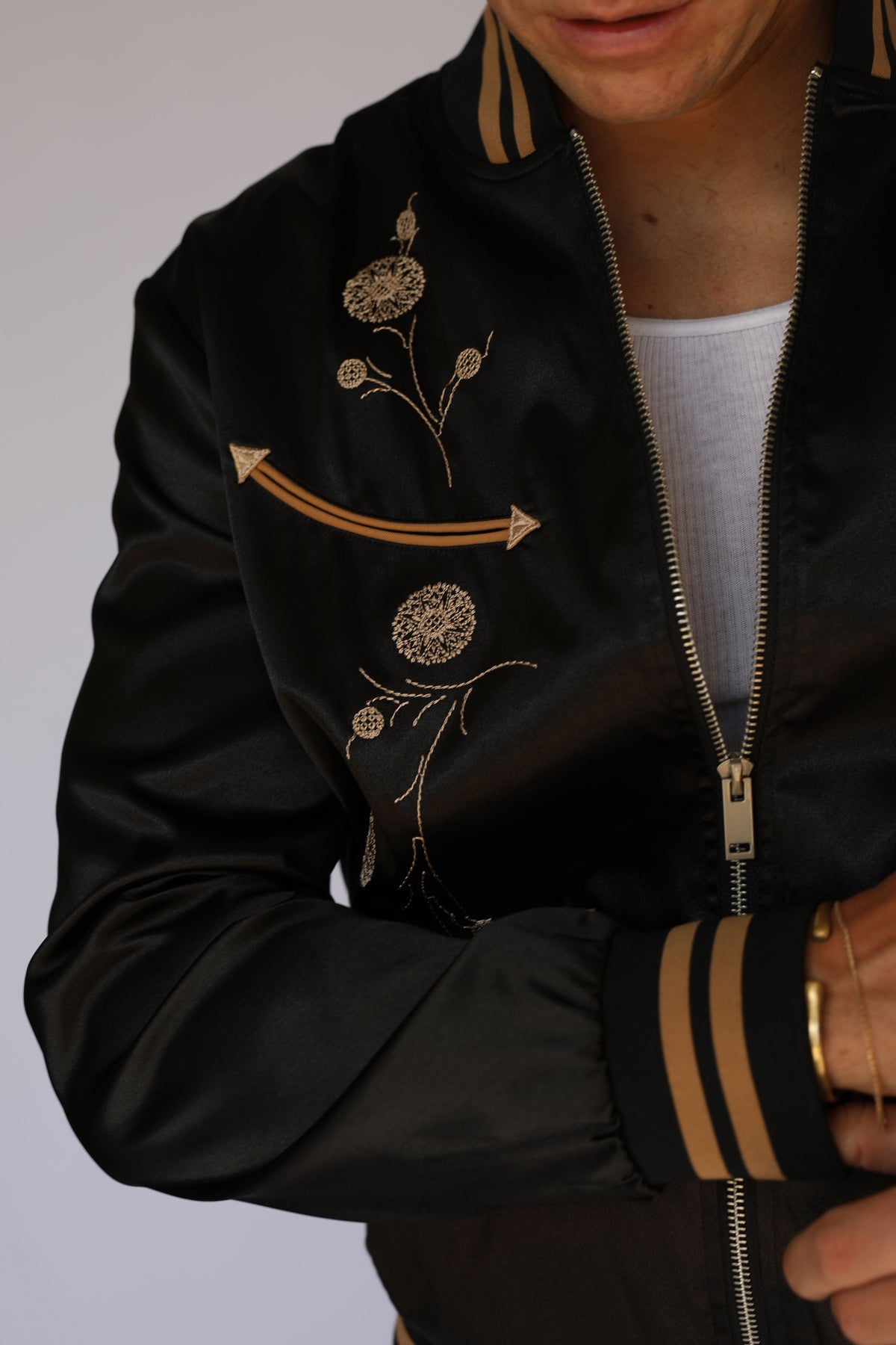 Close-up of the H Bar C x Gibson Odessa black satin jacket showing Gibson J-200 floral embroidery, tan piped arrow pocket, zipper, and striped cuff, vintage western wear detail.