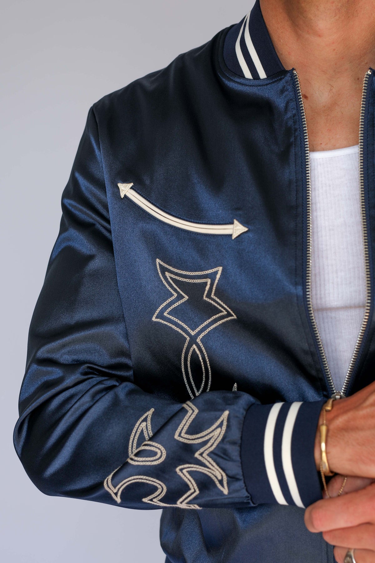 Close-up of the H Bar C Odessa navy satin jacket showing a cream arrow appliqué, chain stitch embroidery, and striped ribbed cuff, vintage western wear detail.