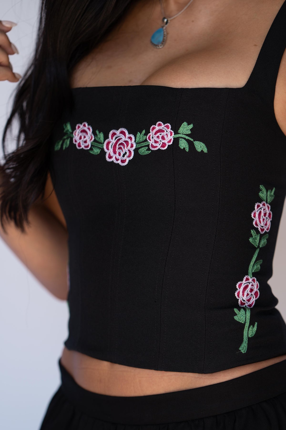 A close-up of a black sleeveless crop top with pink and white embroidered roses and green leaves, worn by a model.
