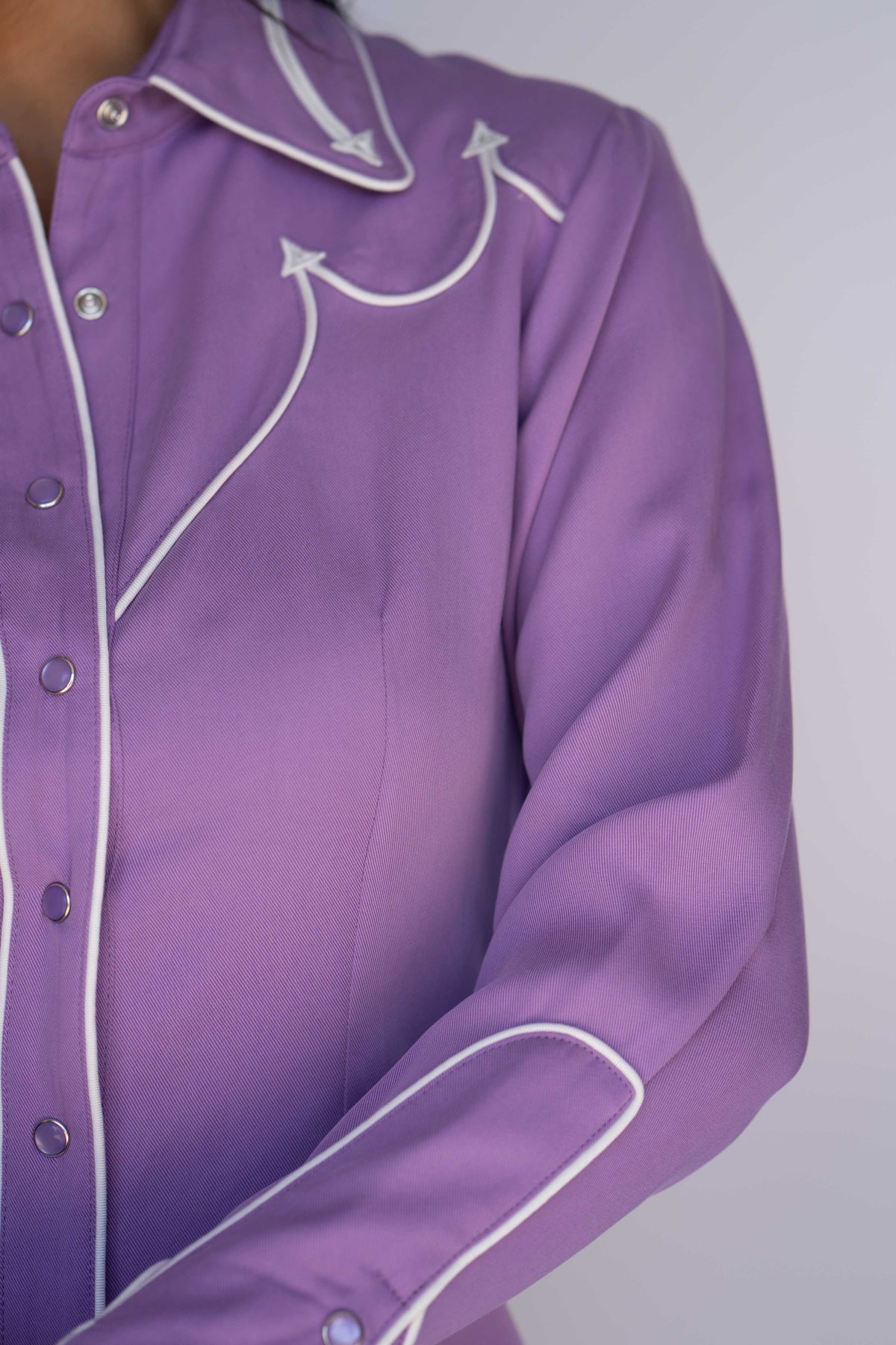A close-up of a lavender purple long-sleeve western-style blouse with white contrast piping and silver buttons, shown on a model against a light gray background.