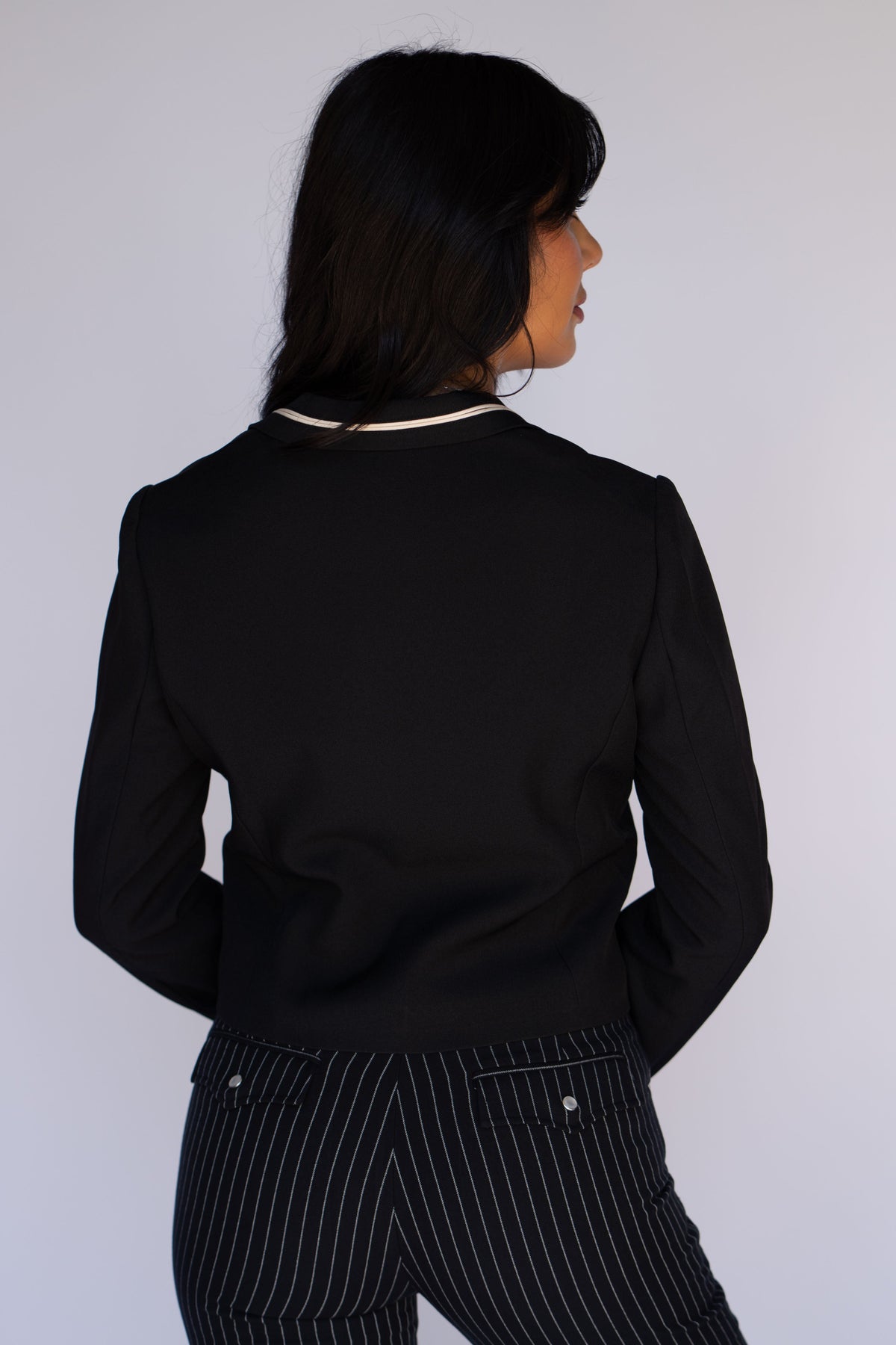 A woman shown from the back wearing a black cropped blazer with a thin cream collar trim and black pinstriped trousers, against a plain light gray background.