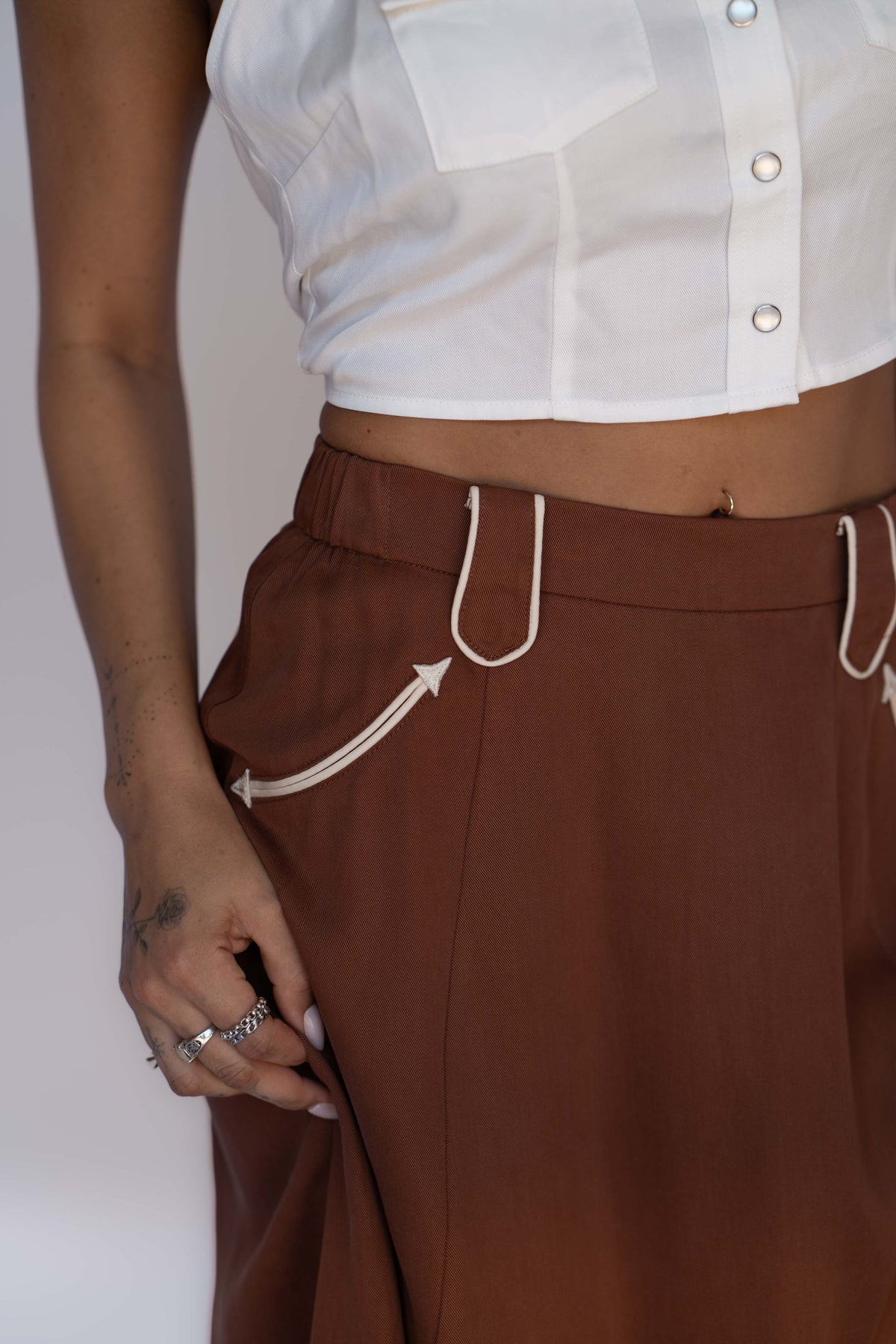 A close-up of a person wearing brown wide-leg trousers with white contrast stitching and a white cropped button-up top.