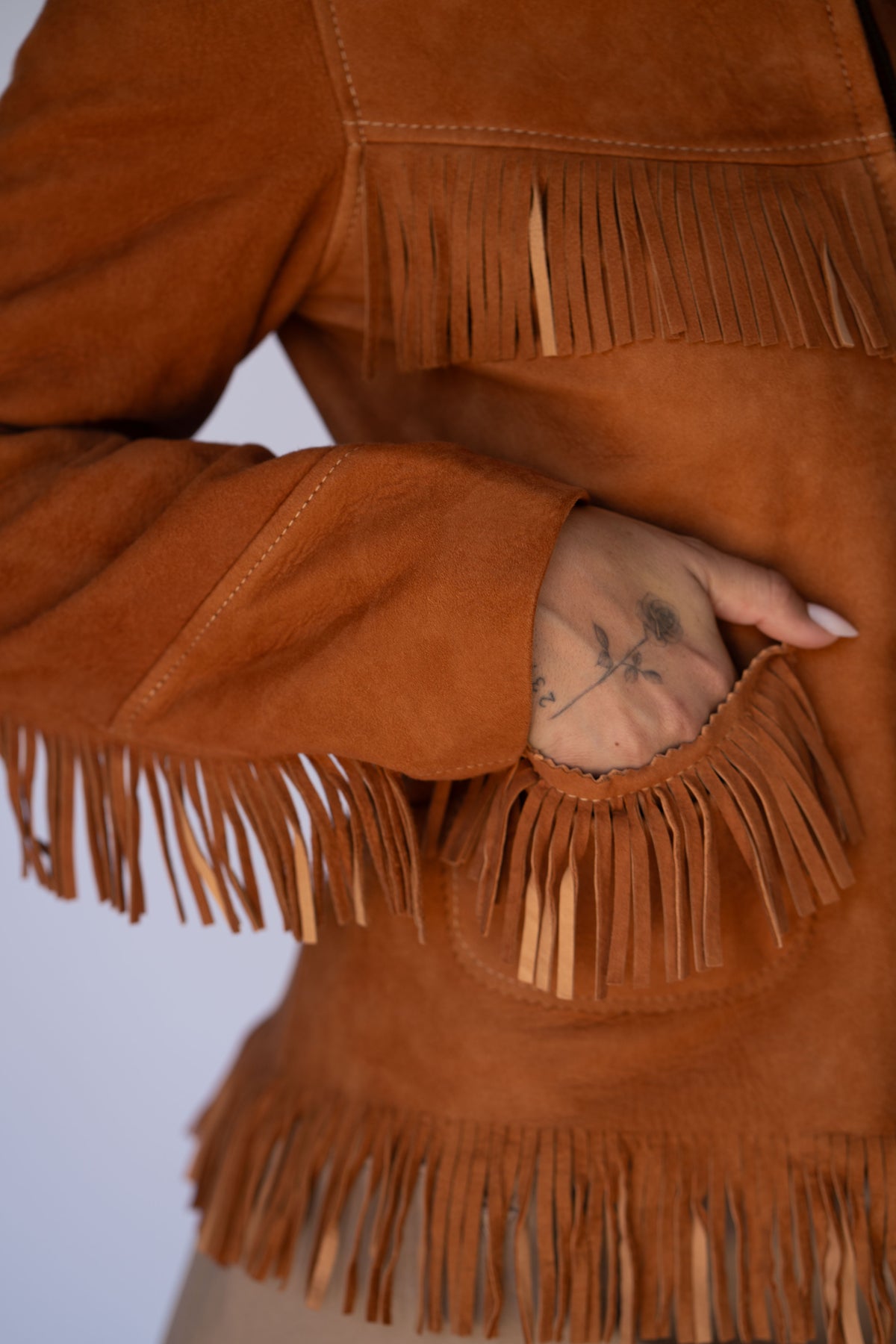 Close-up of a person wearing a rust-brown suede fringe jacket, with one hand tucked into a front pocket.