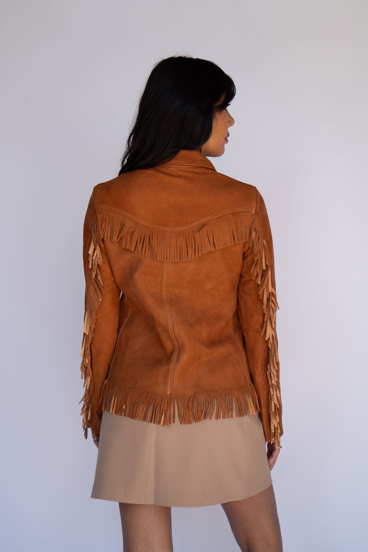 A woman shown from the back wearing a tan suede fringe jacket over a beige skirt, photographed against a plain light gray studio background.