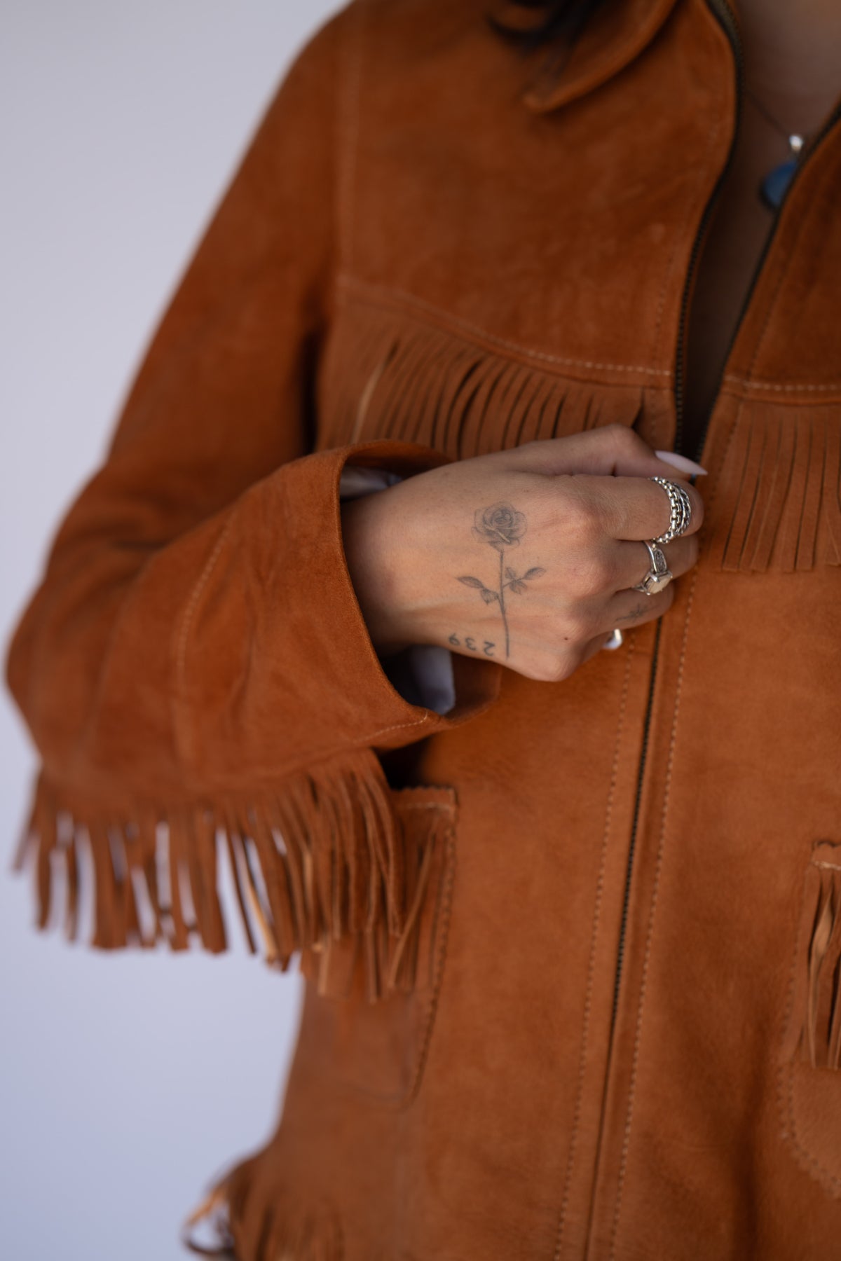 A close-up of a person wearing a rust-brown suede fringe jacket, showing the hand adjusting the zipper. The hand has a small floral tattoo and several silver rings.