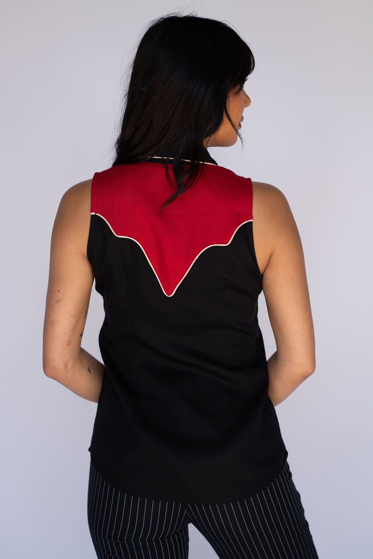 A woman shown from the back wearing a sleeveless black top with a red back panel and white piping, paired with black-and-white pinstriped pants, against a plain light gray background.