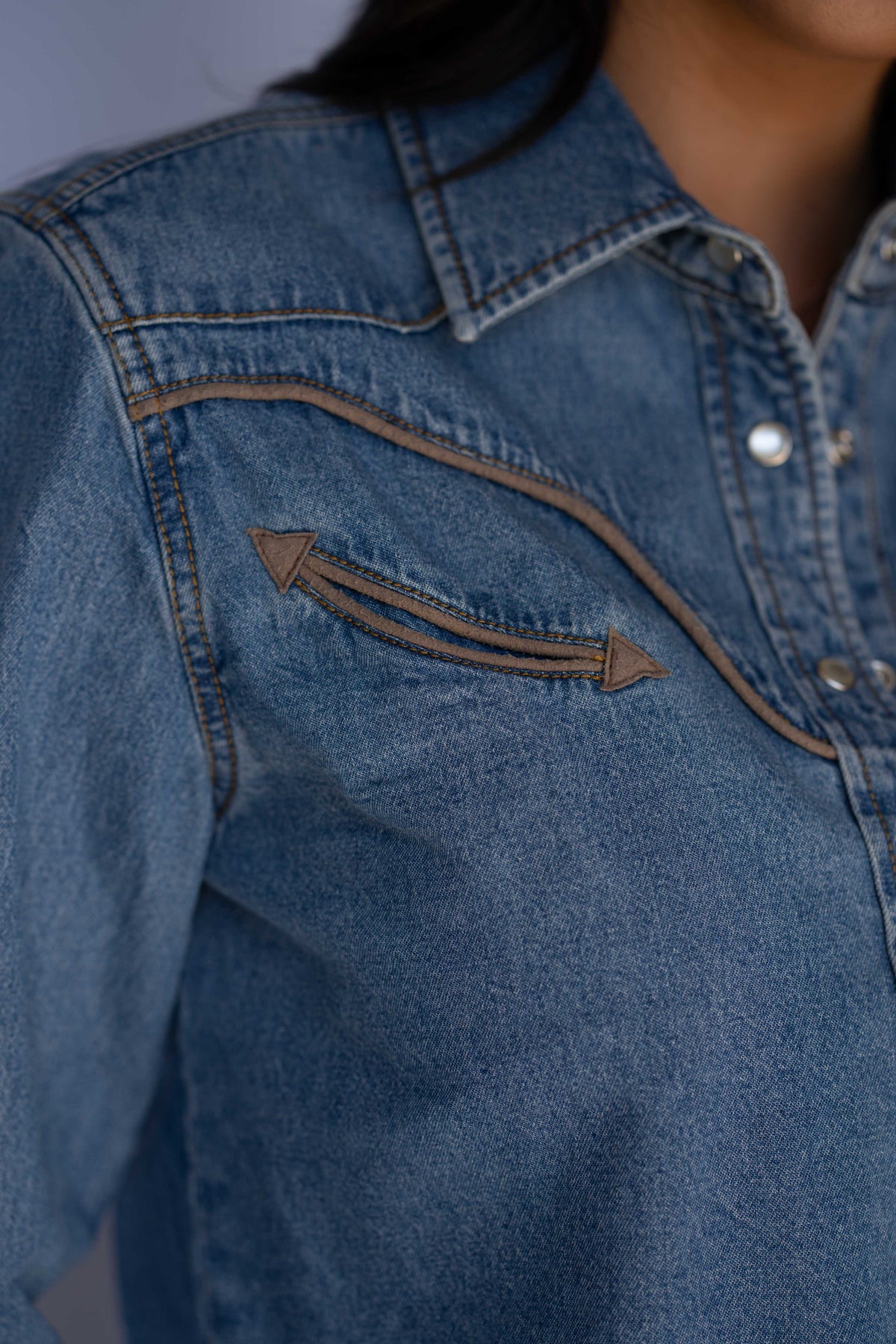 Close-up of a blue denim shirt jacket with a western-style chest pocket featuring double arrow stitching.