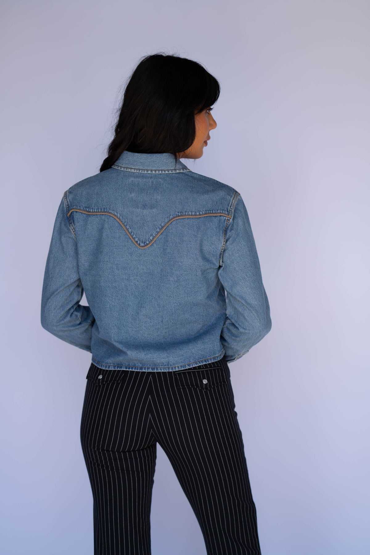 A woman shown from the back wearing a light blue denim jacket with decorative contrast stitching and black pinstriped trousers, standing against a plain light gray background.