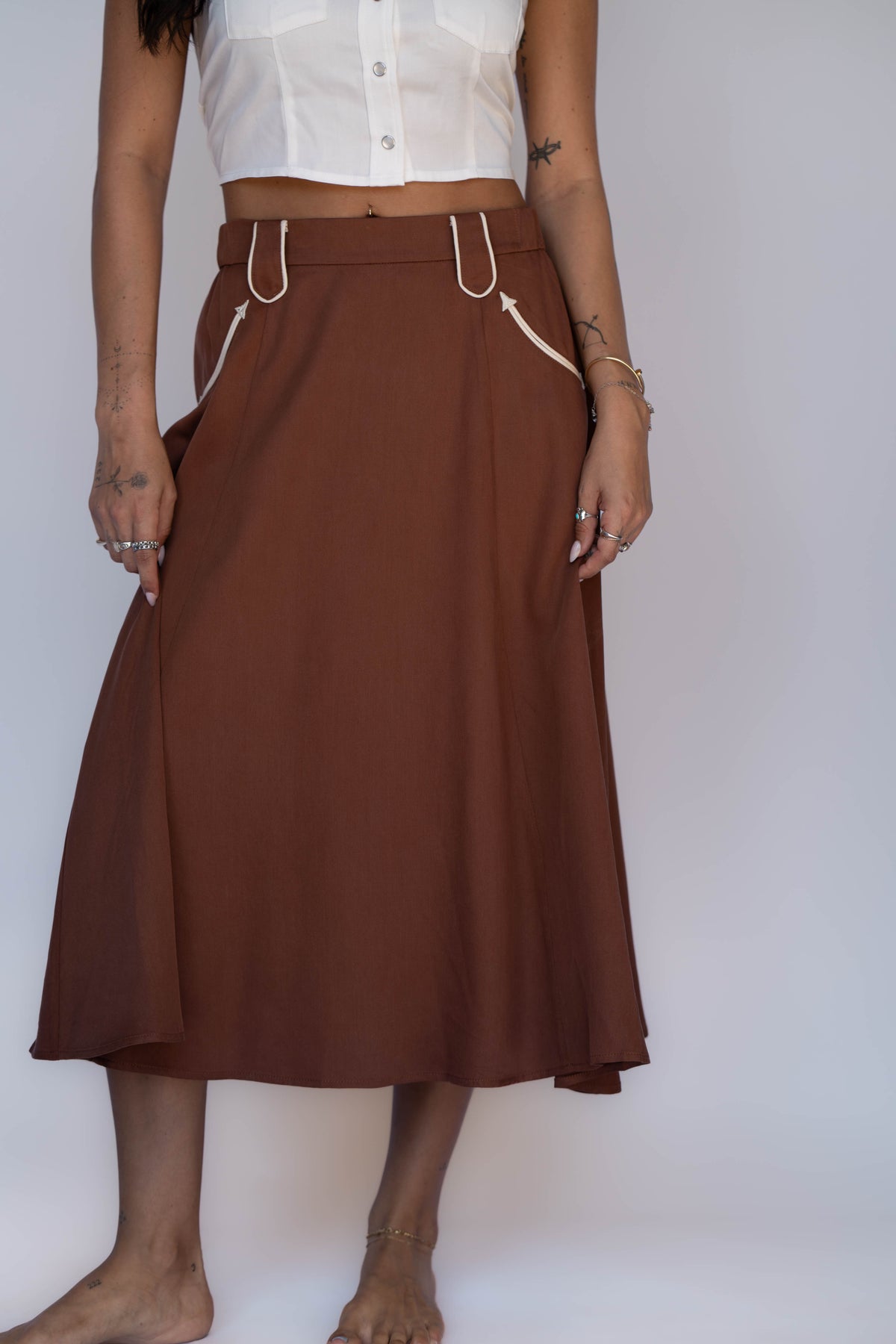 A woman wearing a brown A-line midi skirt with white drawstring details at the waist and side pockets, paired with a white cropped top, standing against a plain light gray background.