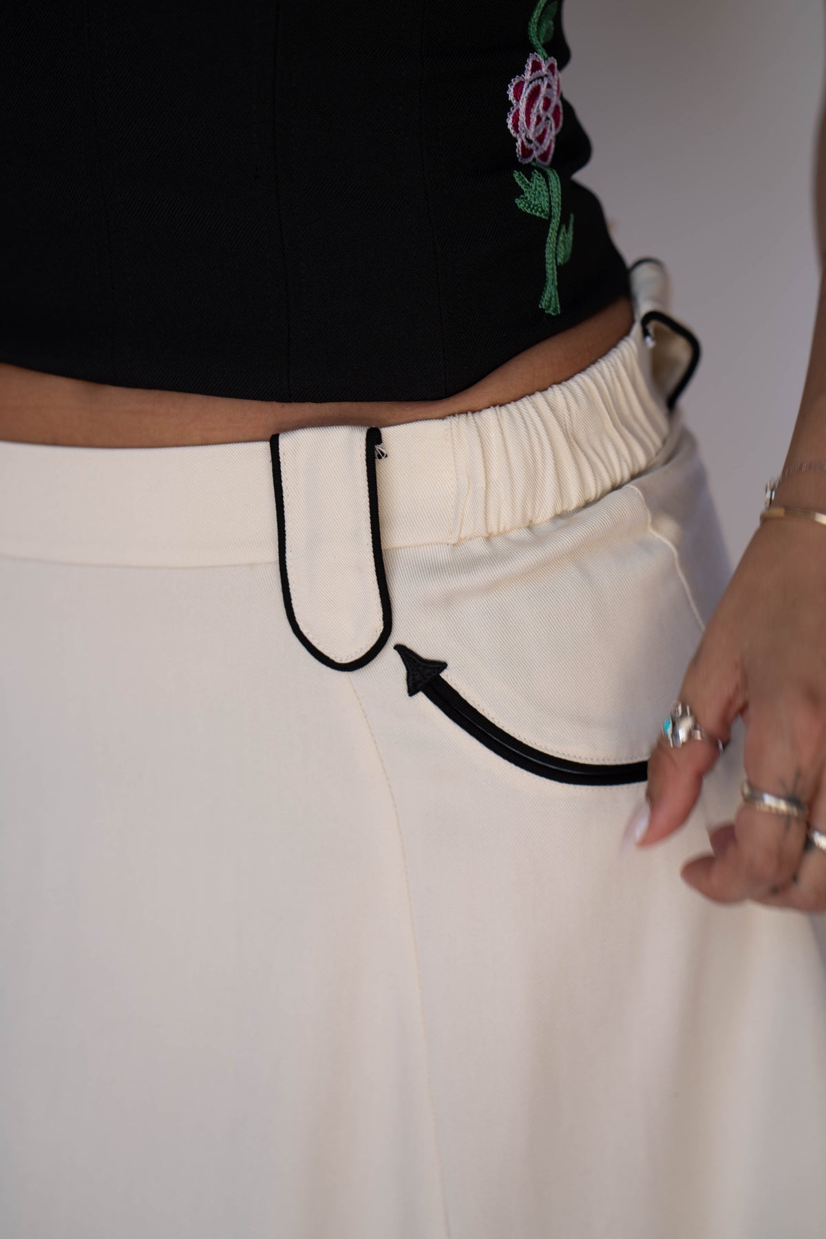 Close-up of a person wearing off-white trousers with a distinctive black curved hook detail at the front waistband and a black drawstring, paired with a black cropped top.