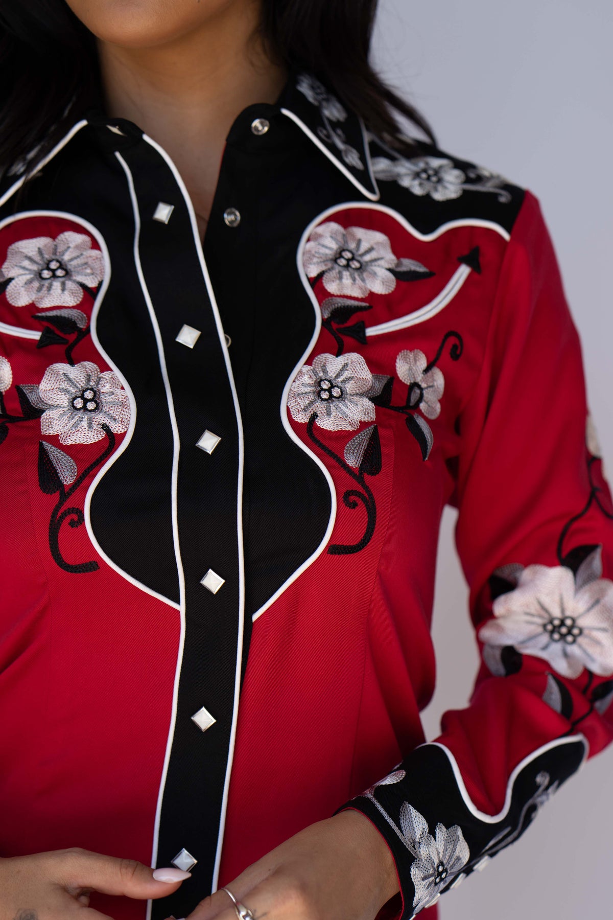 A close-up of a red long-sleeve western-style blouse with black yoke panels, white embroidered floral motifs, and white piping details.