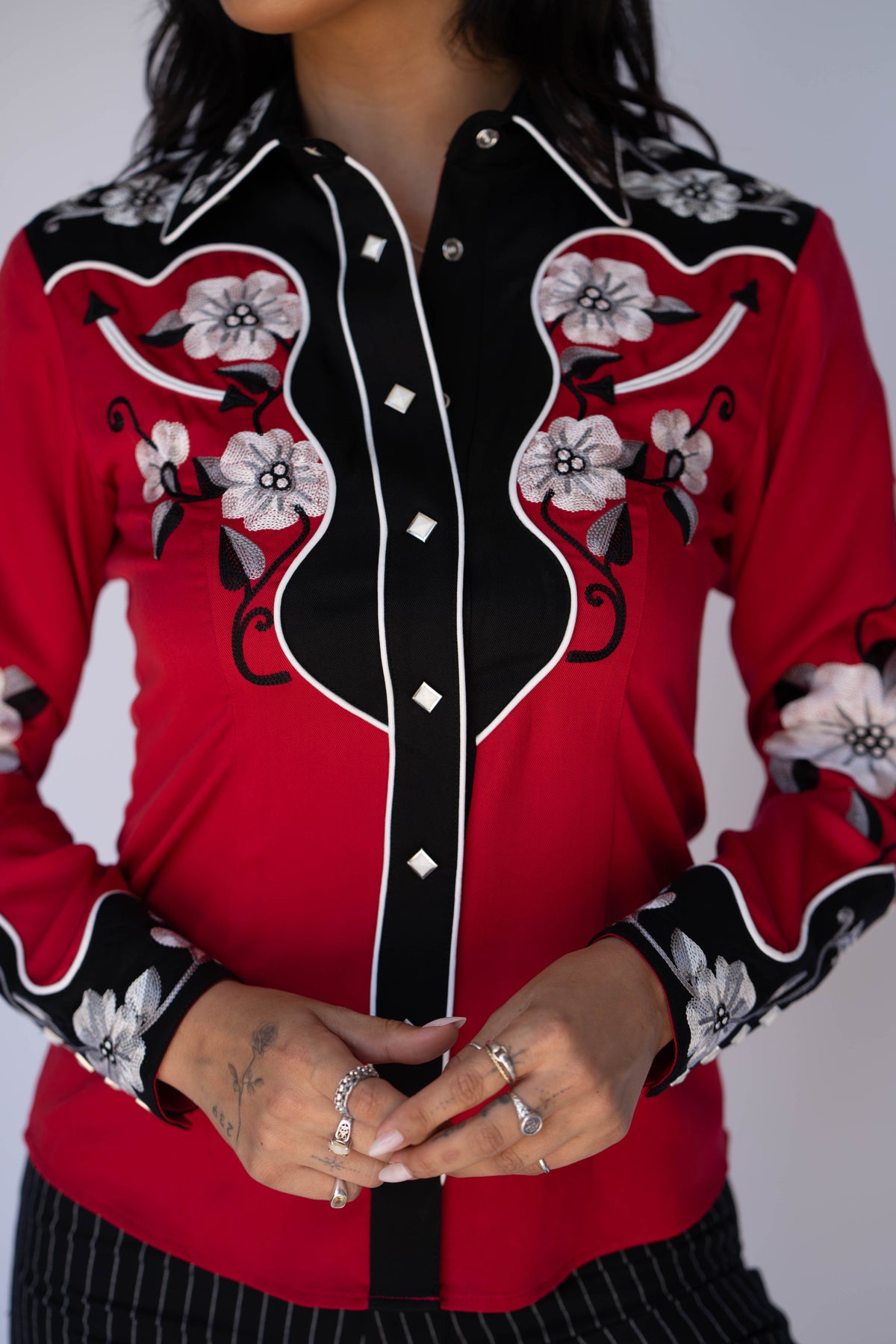 A close-up of a red western-style long-sleeve button-up shirt with black yoke panels, white floral embroidery, and white piping details, worn by a model.