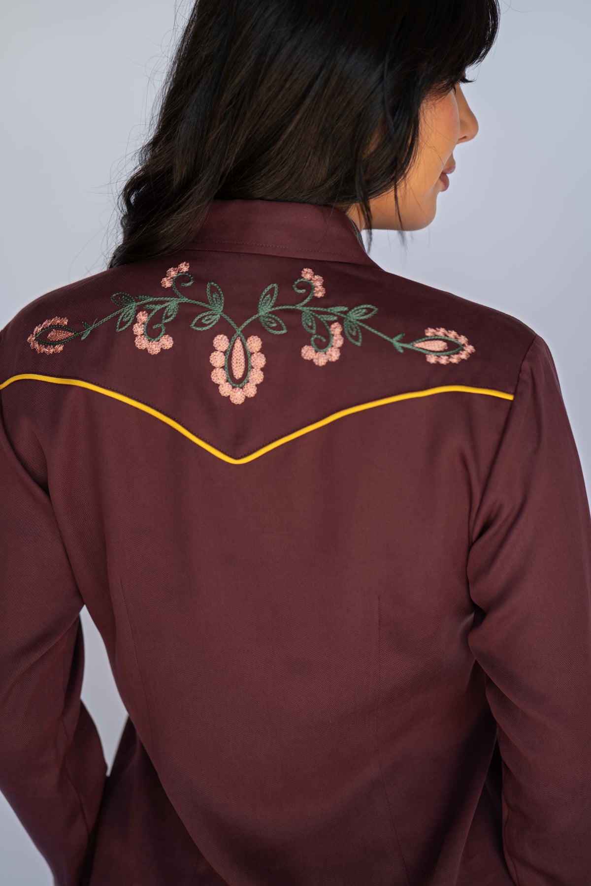 A woman shown from the back wearing a dark brown western-style shirt with floral embroidery across the shoulders and yellow piping along the yoke.