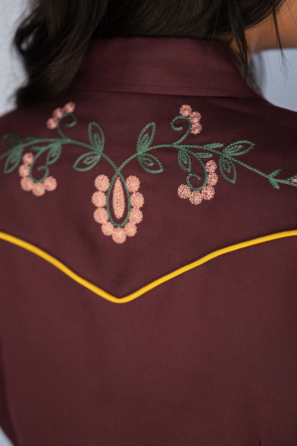 Close-up of the back of a burgundy western-style shirt with green and pink floral embroidery across the shoulders and a yellow contrast stitch along the yoke.