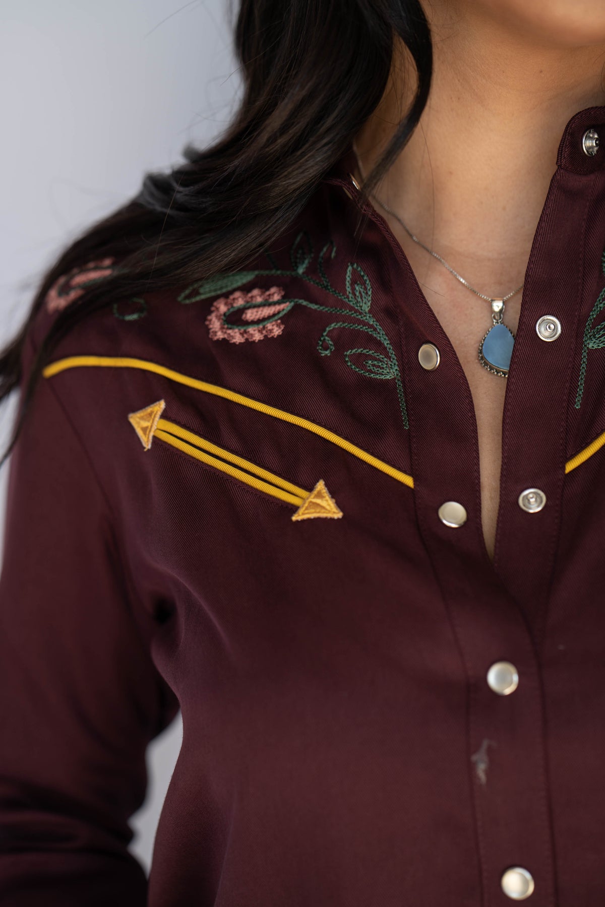 Close-up of a woman wearing a burgundy western-style button-up shirt with embroidered floral details, yellow piping, and gold-toned snap buttons.