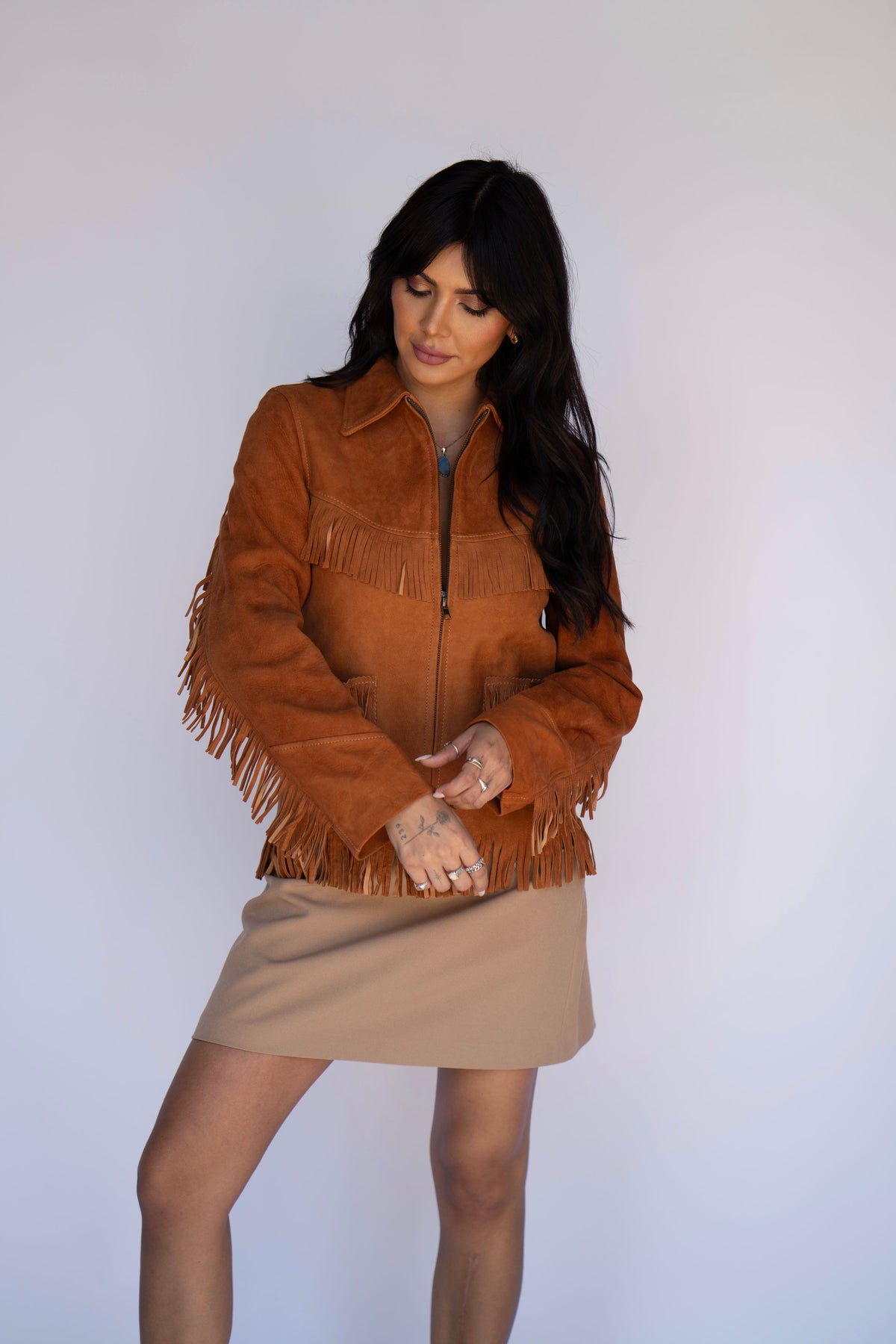 A woman modeling a brown suede fringe jacket over a beige mini skirt, standing against a plain light gray studio background.