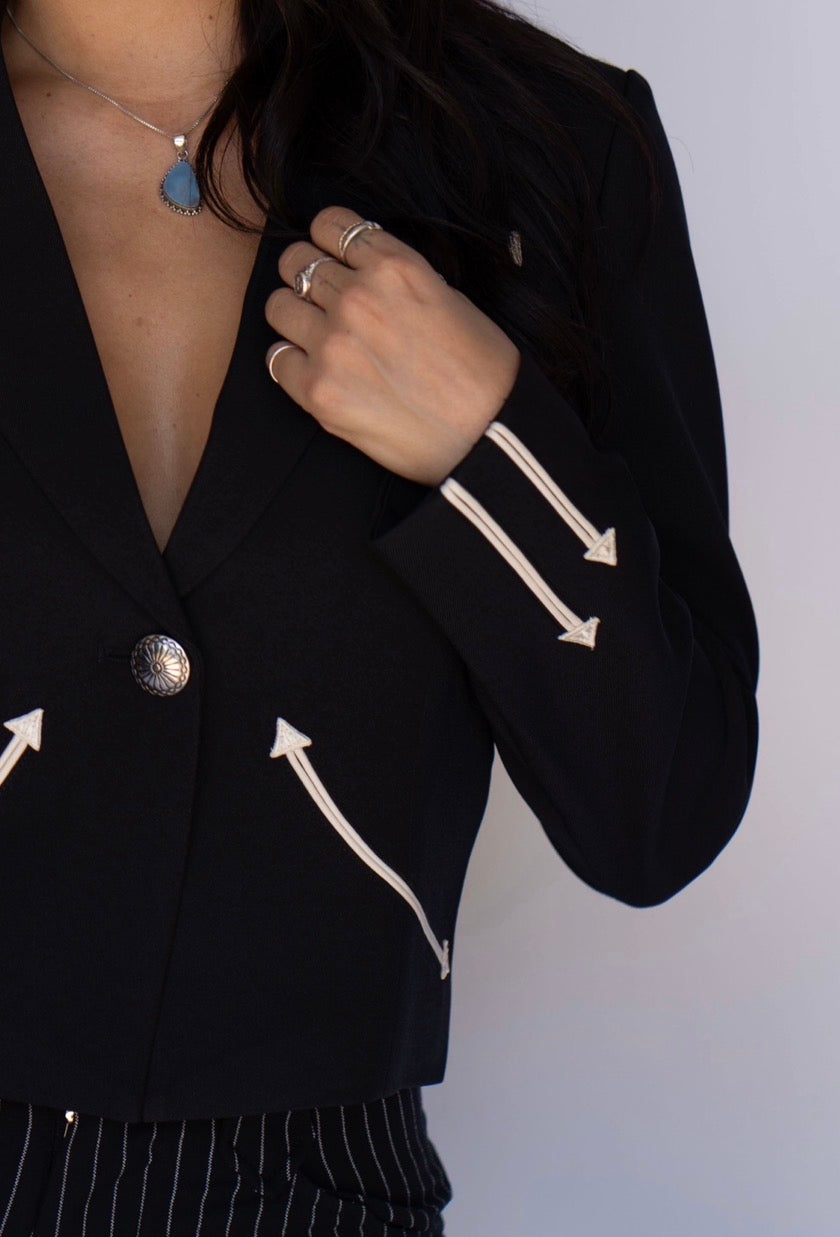 A close-up of a person wearing a black blazer with cream-colored embroidered arrow details on the sleeves and front, paired with striped trousers and a necklace.